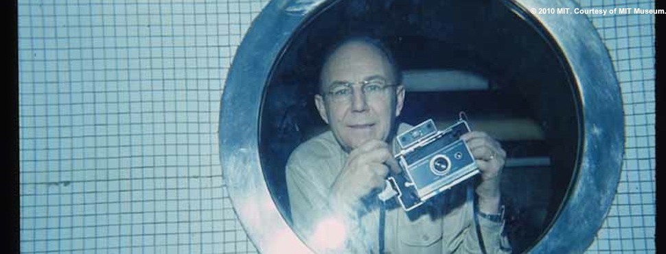 H. E. Edgerton holds a (Polaroid?) camera, photographed from inside the MIT pool, as he peers through an underwater window.